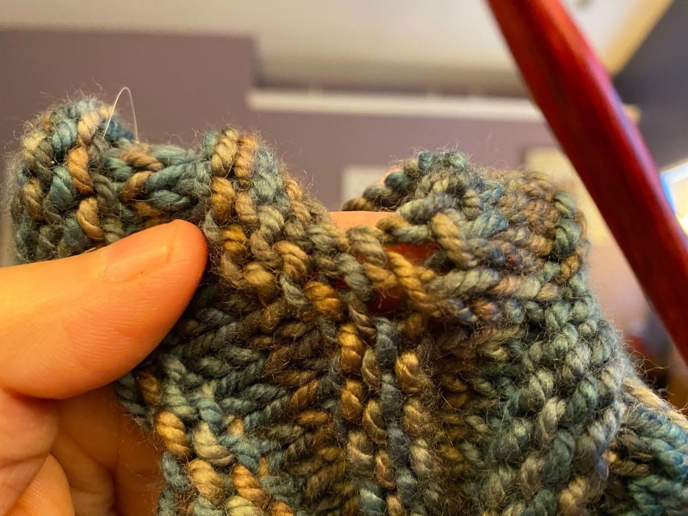 A knitted cowl in chunky green and gold yarn. Two obvious holes shown.