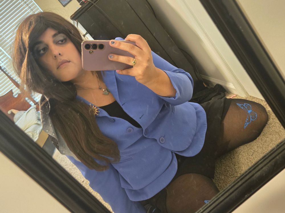Ira kneeling in front of a mirror and taking a selfie, wearing her hair in a ponytail with a Kingdom Hearts themed bow. She is wearing a blue blazer with a black tank top underneath, a black pencil skirt, and black tights with blue butterflies patterned onto them. Her hair is a bit messy in the photo.