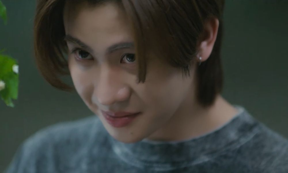 a imagem foca no rosto e parte do busto de um rapaz tailandês de cabelos castanhos e partidos ao lado da testa. ele está olhando para cima com a cabeça um pouco inclinada para baixo, com um pequeno sorriso no rosto. o fundo da imagem é verde escuro e no canto esquerdo há um pedaço de planta aparecendo desfocado