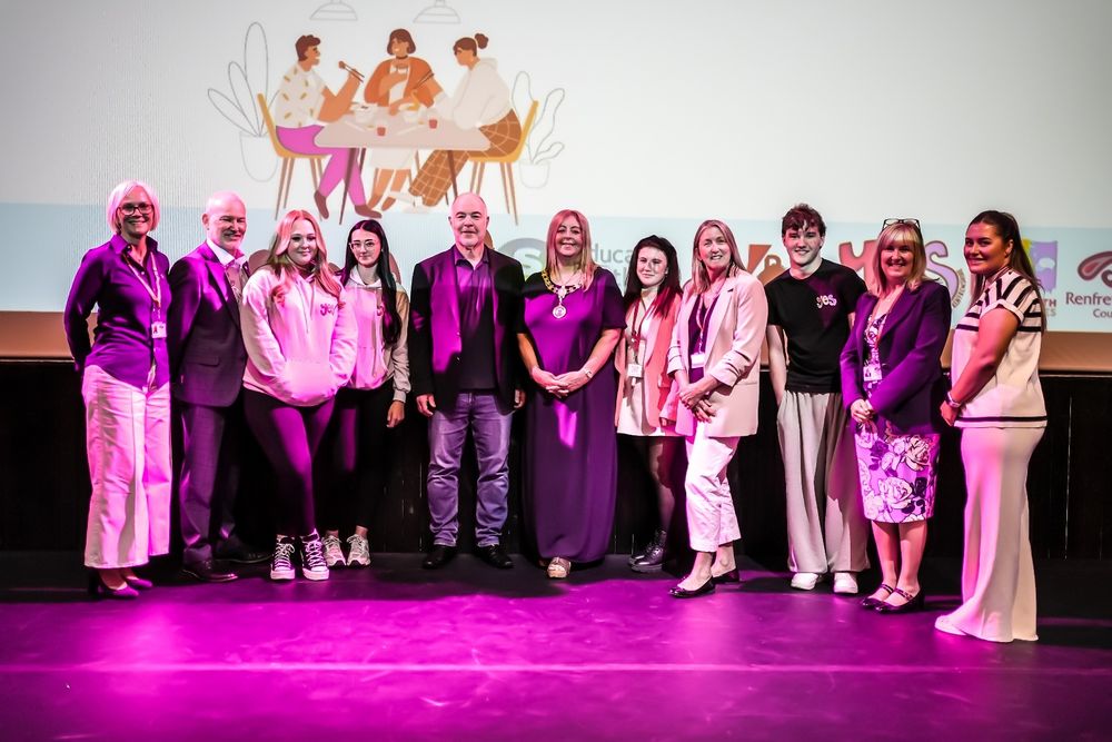 A group of young people and adults stand on a stage in a row under purple lighting.