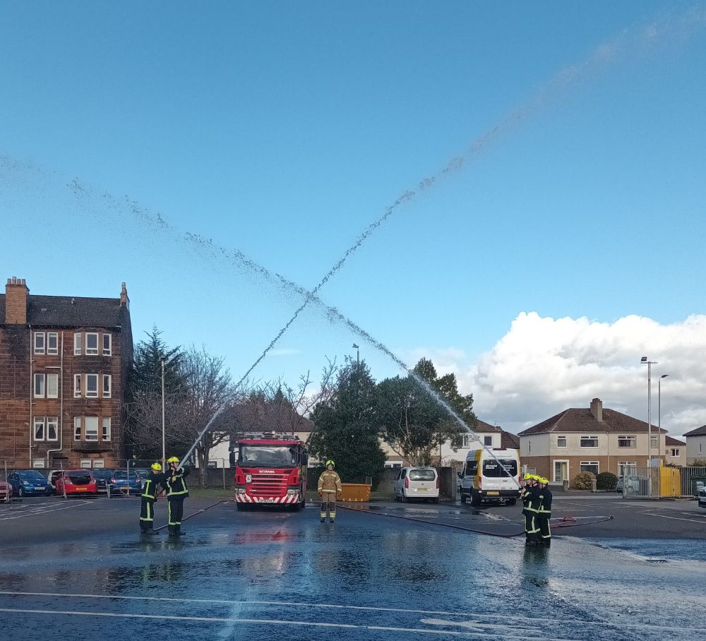Young people wearing fire safety gear stand in two groups spraying water from hoses into the air.
