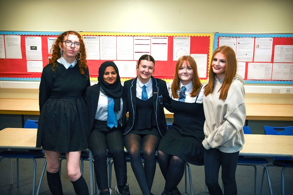 Five young people sit in a row on classroom desks and smile.
