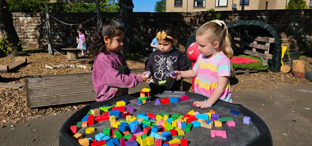 Three young children play with coloured blocks at a table outside in the sunshine.