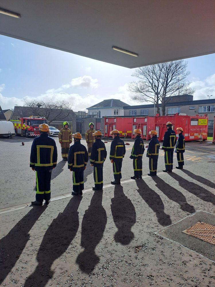Young people wearing fire safety gear stand outside listening to a fire officer explain fire safety.