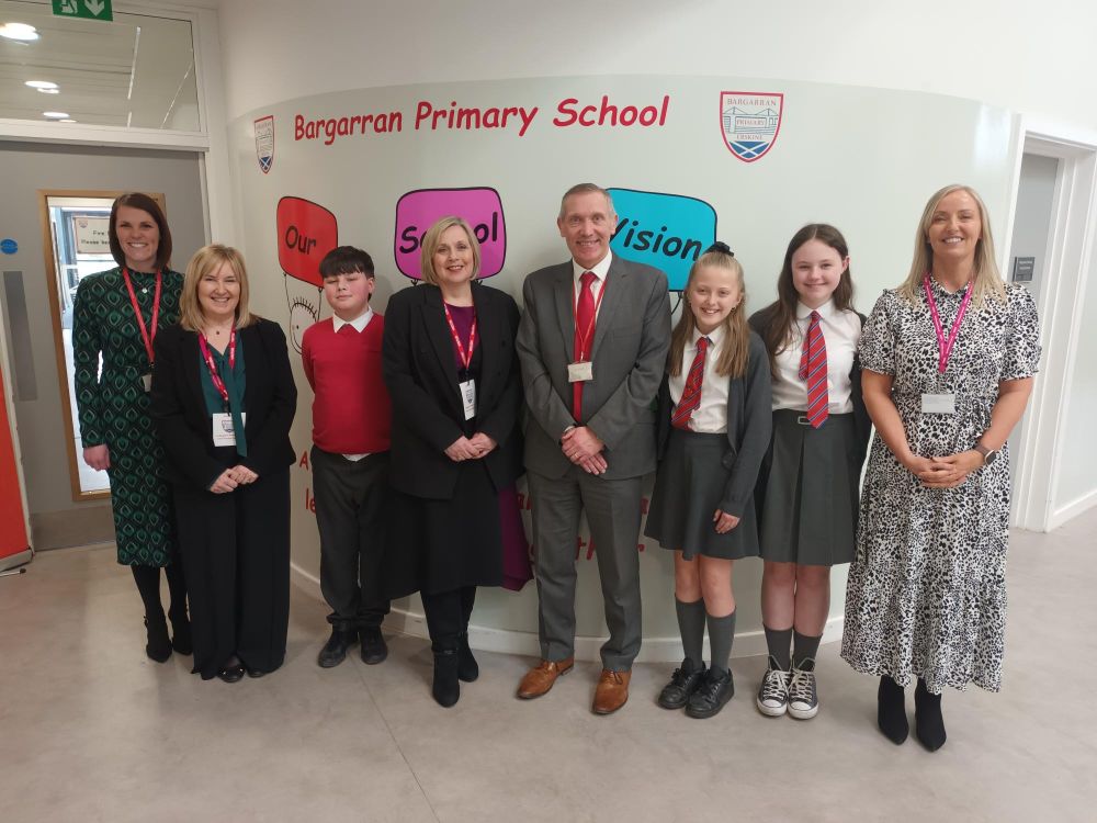 Janie McManus, Janie O'Neill, Alistair Lyle, Jennifer Thompson, Selena Kennedy and primary seven pupils Connor, Imogen and Charlie standing in front of a Bargarran Primary School sign within the school.