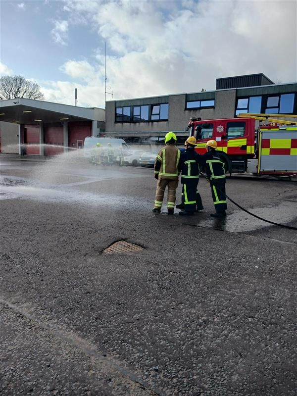 Young people and a fire officer wearing fire safety gear and using a hose to spray water.