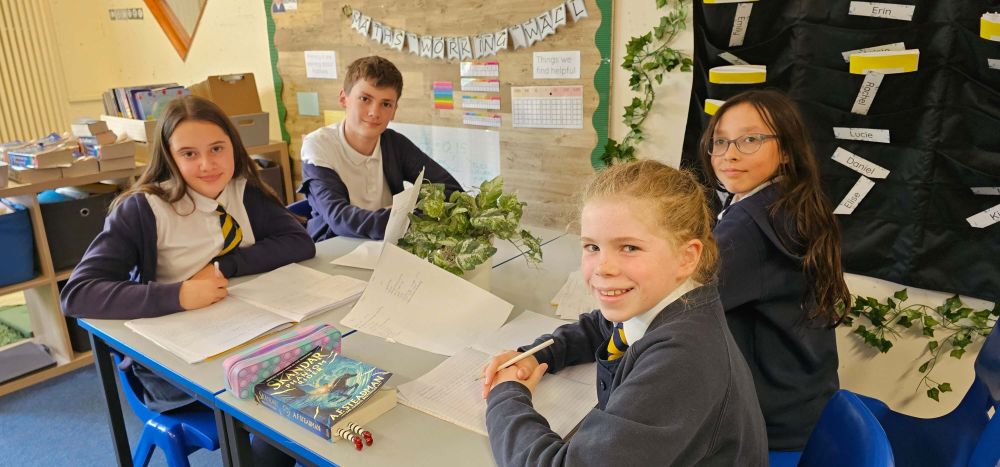 Four pupils smile while seated around a desk doing coursework.