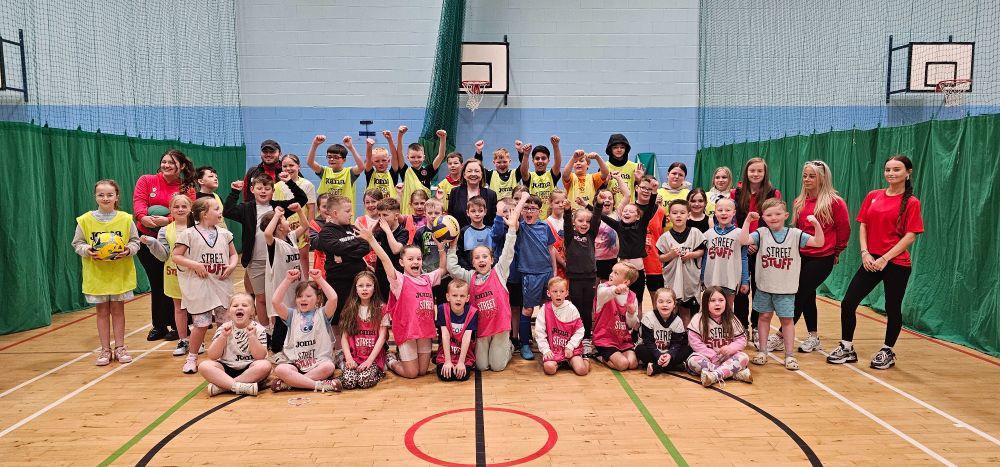 A large group of children and adults gather together in a gym hall.