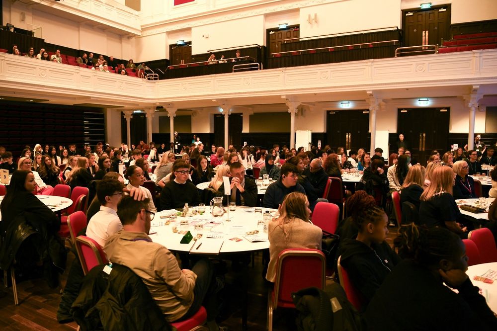 Hundreds of young people sit at round tables in a large room.