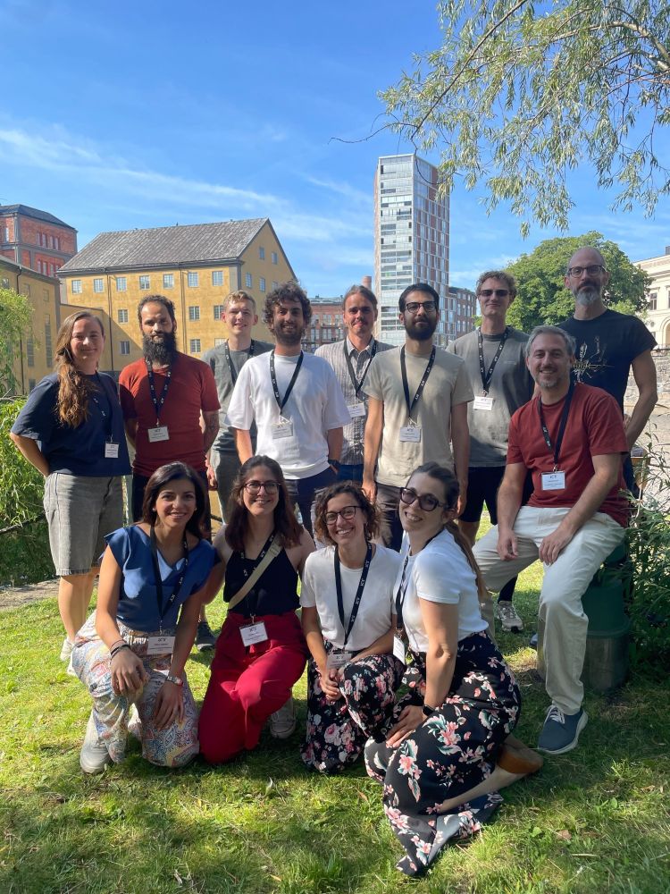 Photo of a group of young researchers smiling, colorful clothes, on grass, historical industrial background of Norrköping
