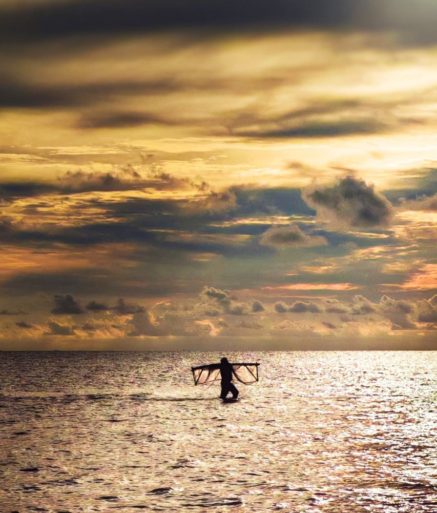 A silhouette of a person walking through shallow ocean water at sunset, carrying a large fishing net over their shoulders, with golden light reflecting on the water and dramatic clouds filling the sky.