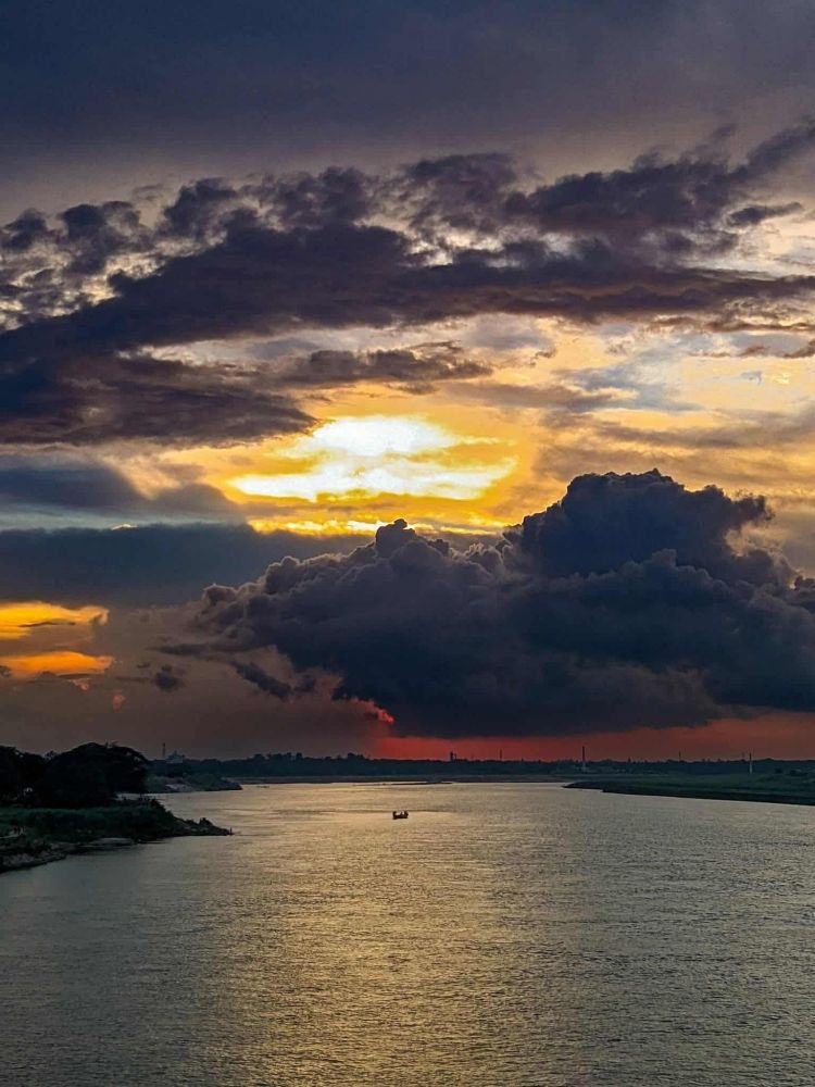 A stunning sunset scene with dramatic dark clouds and golden-orange light reflecting off a calm river. A small boat floats quietly in the center of the frame, surrounded by peaceful nature.