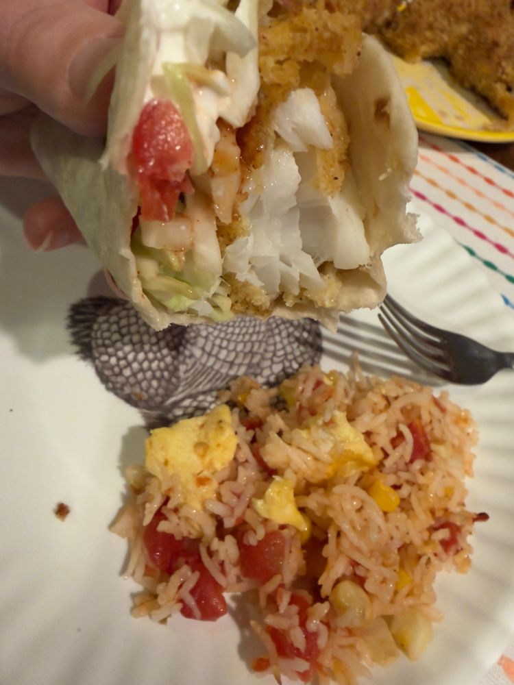 A hand holding a taco in a flour tortilla. There is a bite taken, showing the flaky, white cod. On the plate below, there is rice with chunks of tomato and scrambled egg. 