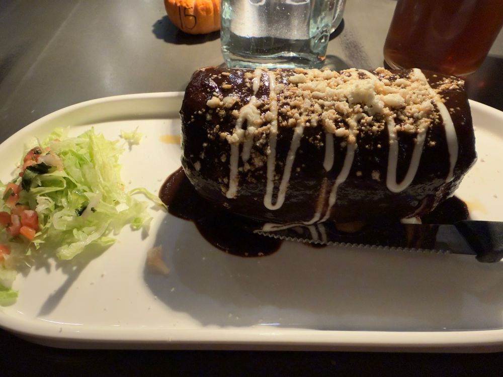 A large chimichanga covered in a very dark mole sauce with ribbons of crema and cotija cheese sprinkled on top. 