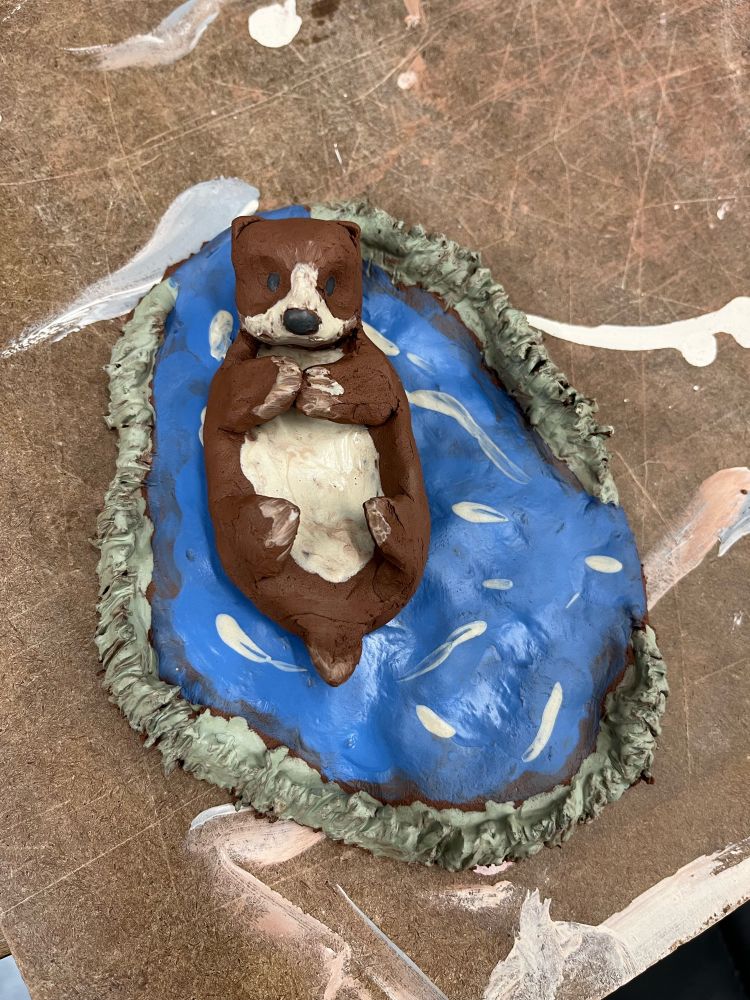 Image of an otter on its back, which is made out of brown clay. It is sitting on another base of clay that looks like water. (Or so I hope.) 