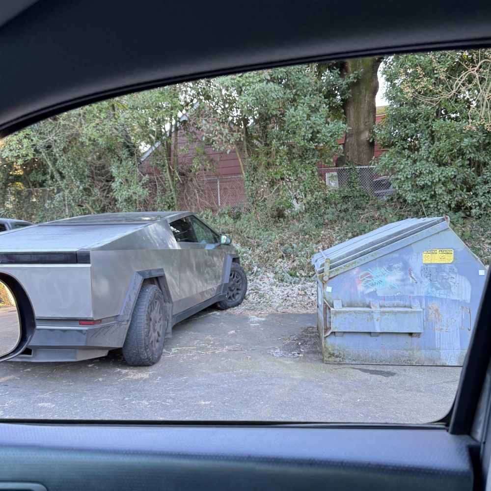 An image of a boxy angled silver dumpster (left) next to another dumpster (right).