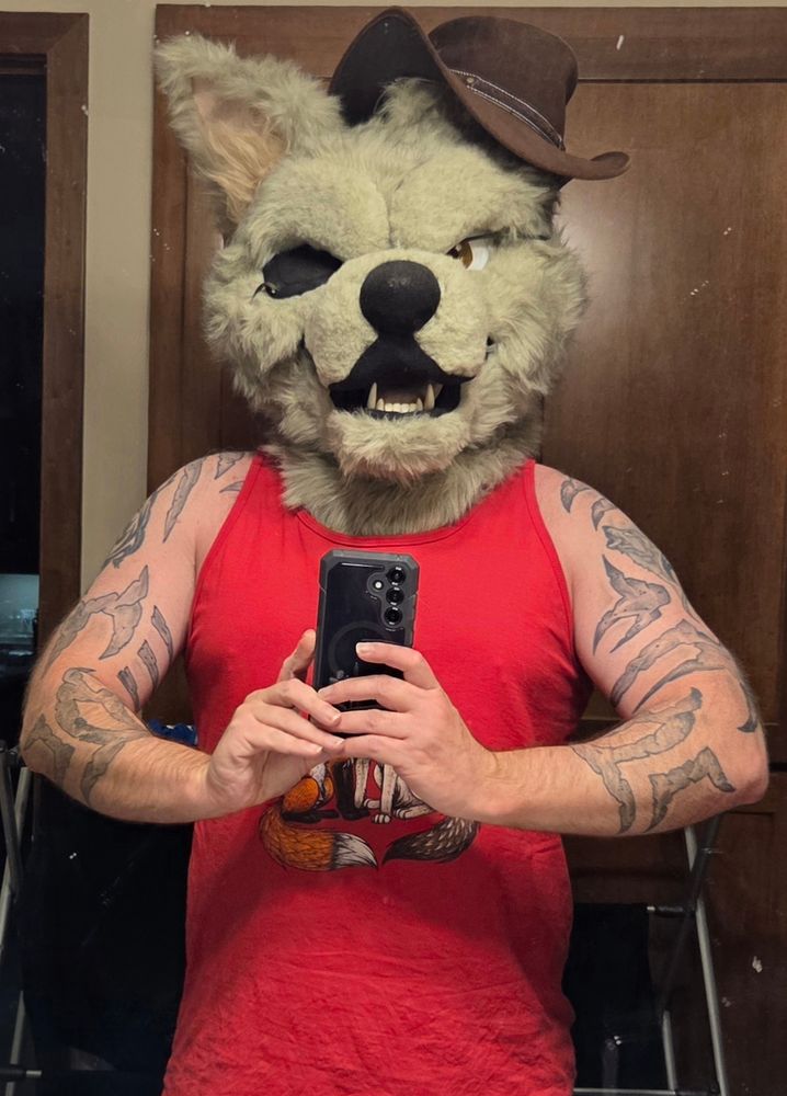A grey wolf fursuiter wearing a red sleeveless shirt. They are taking a selfie in the mirror.