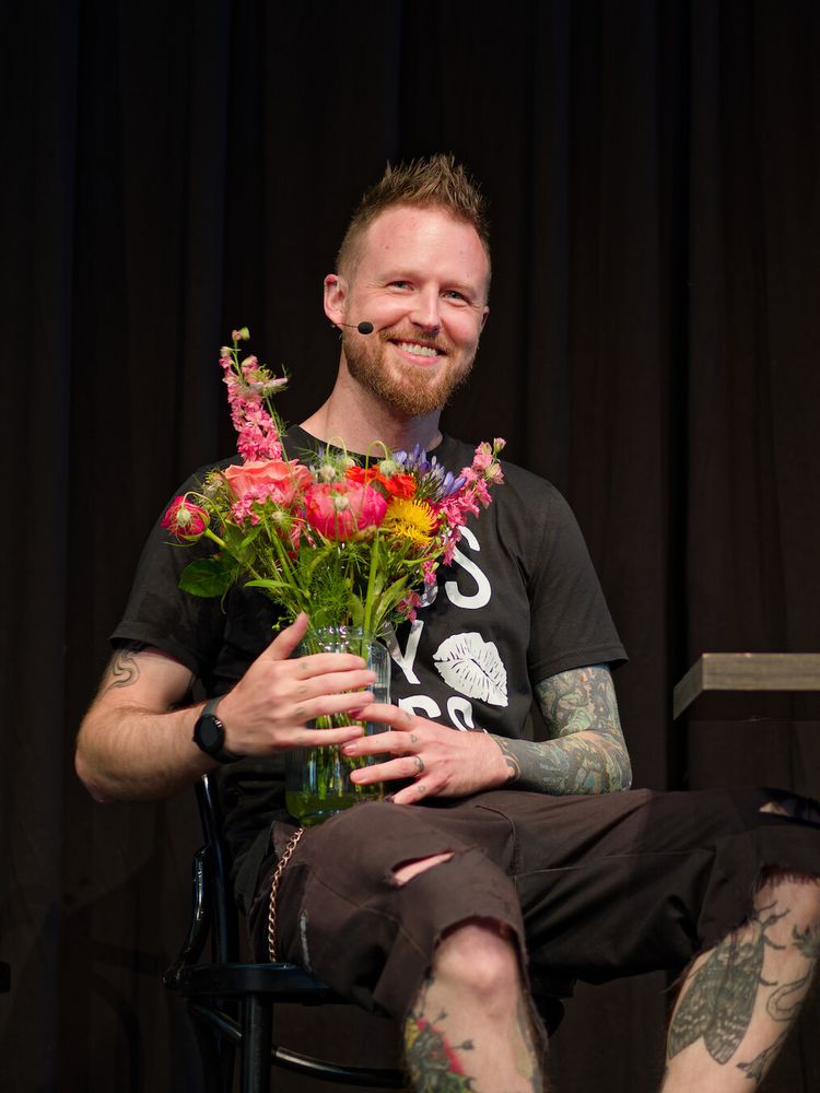 A photo of Adam sitting on a chair, holding a vase with various colorful flowers and smiling. There are more tattoos visible on Adam's arms and legs.
