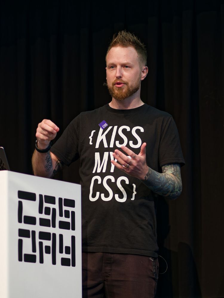 A photo of Adam giving a talk and gesticulating near a podium while looking at the audience. A full “CSS Day” logo is now visible on the podium, and a full “Kiss My CSS” print on Adam's t-shirt, with curly brackets around this text.