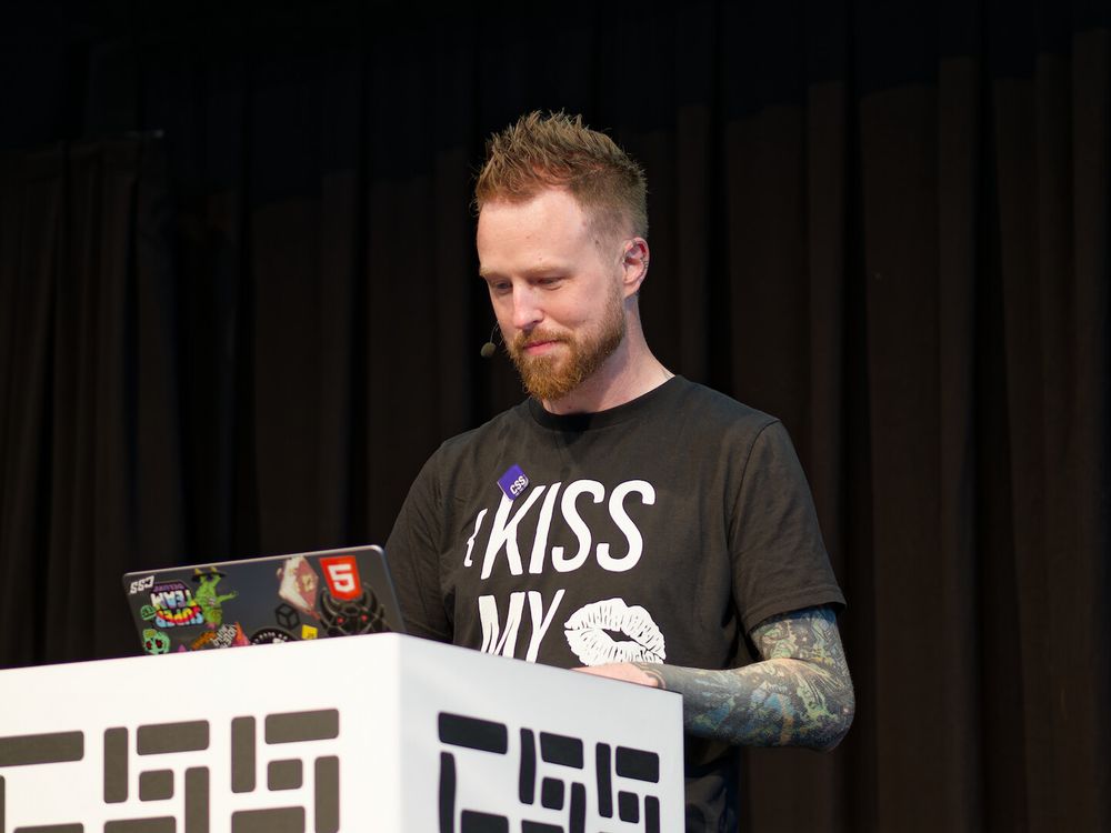 A photo of Adam standing near a podium with “CSS” visible on it and an open MacBook with various stickers on its lid. Adam is looking at the MacBook's display and is wearing a black t-shirt, with something starting from “Kiss My” on it, followed by lips. There is a purple “CSS logo” pin on the t-shirt. A full sleeve tattoo is visible on Adam's left hand.