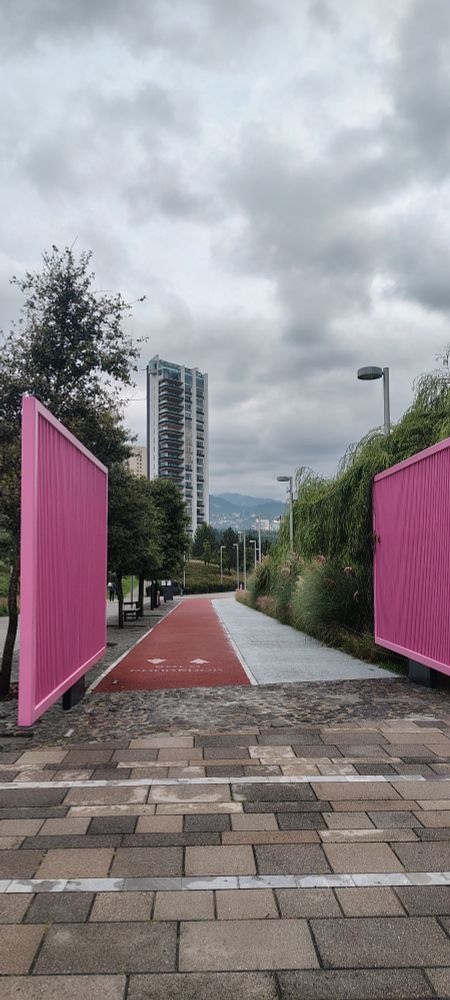 Two large pink gates frame the entrance to a red running track in the park, with tall grass, trees, and a modern high-rise apartment building in the background