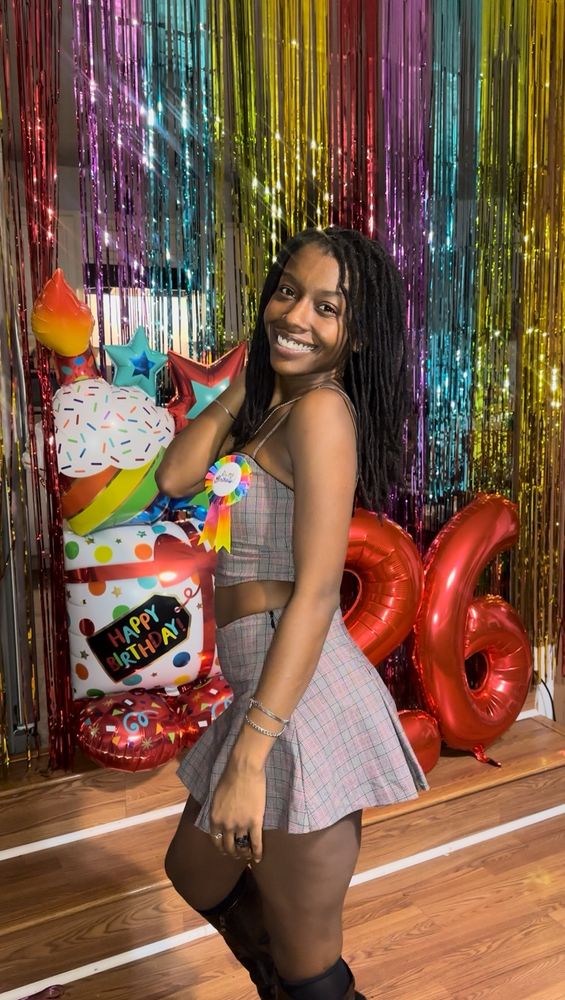 Marley is smiling in front of a colorful backdrop and birthday balloons. She facing to the side with one hand by her side and the other hand on her locs. She is wearing a gray, red and black plaid two piece.