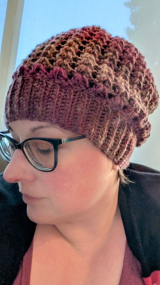 Woman with glasses wearing a handmade slouchy beanie in a burgundy/brown variegated acrylic and wool yarn. 