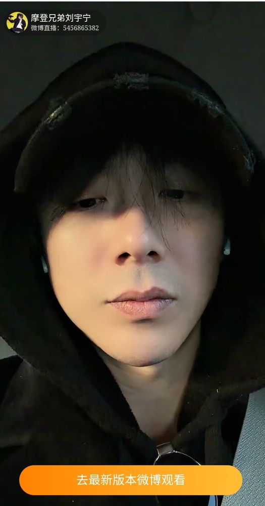 Liu Yuning sério, olhando pra baixo, só o rosto bem perto do celular. Ele está de boné e capuz preto e sem maquiagem, só filtro de base do celular.