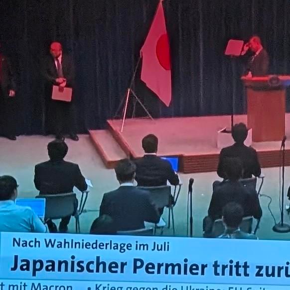 Pressekonferenz mit japanischer Flagge auf der Bühne. Ein Redner steht am Pult, mehrere Personen sitzen im Publikum. Am unteren Bildrand blendet ein Nachrichtensender die Schlagzeile ein: „Nach Wahlniederlage im Juli – Japanischer Permier tritt zur…“.
