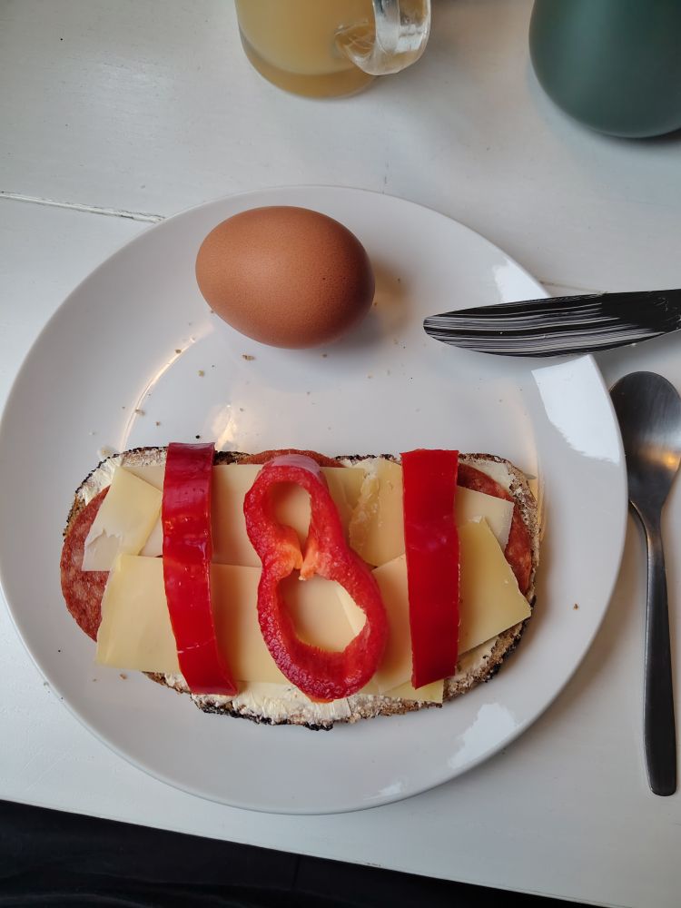 Auf einem weißen frühstücksteller liegt ein braunes gekochtes ei und eine Stulle. Auf dieser ist vegane Salami, etwas Käse und Paprika. Die Paprika ist so geschnitten, dass eine 181 zu erkennen ist. Neben dem Teller liegt ein Löffel. Auf dem Teller ein Messer mit Margarineresten.