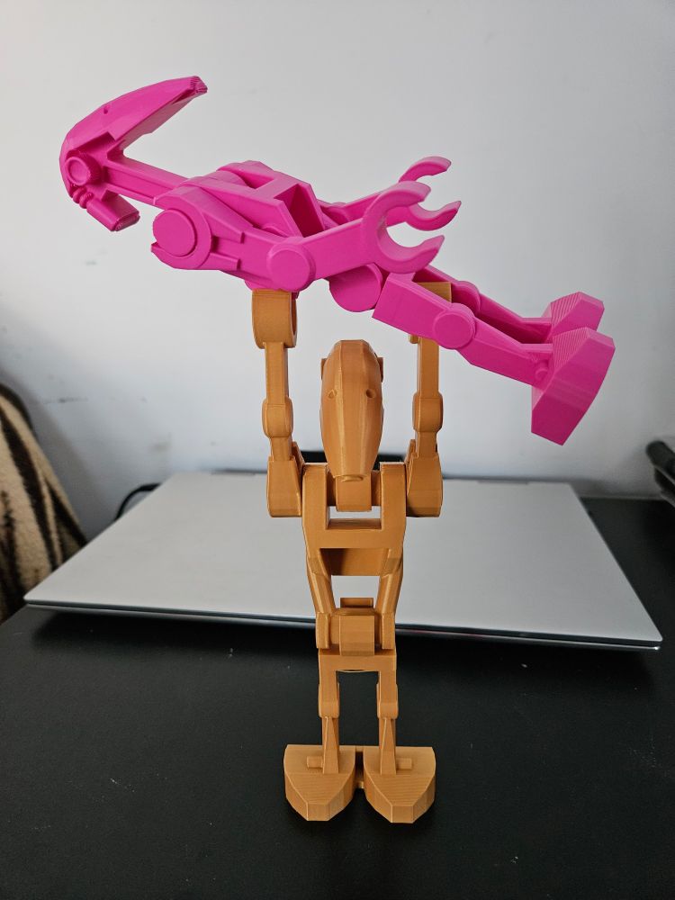A photo of a gold-coloured 3D printed LEGO Battle Droid from Star Wars holding a pink-coloured 3D printed LEGO Battle Froid from Star Wars. Both droids are much larger than their original LEGO counterpart.