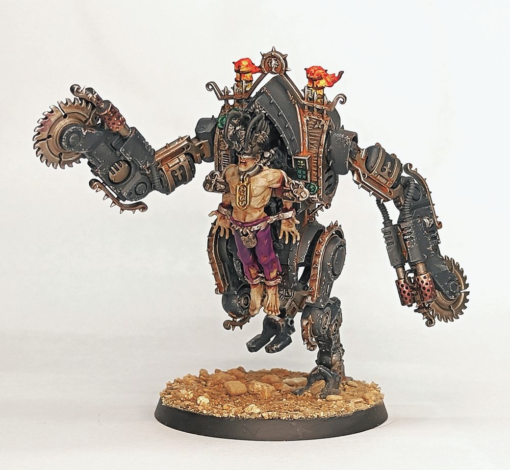 A penitent engine for warhammer 40k. It's arms end in combination circular saws and flamethrowers, as it runs across a desert base. The man strapped to the front of it is wearing purple trousers, but is otherwise having a terrible time. Flames belch from exhausts at the top of the machine. 