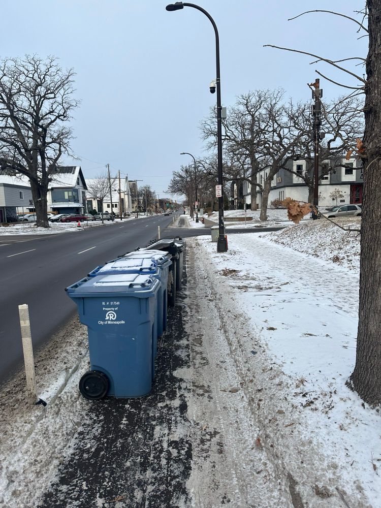 Garbage cans parked in bike lane on SE Fourth Street in Minneapolis. Three car lanes completely clear of snow, while bike lane has a good amount of snow in it. 