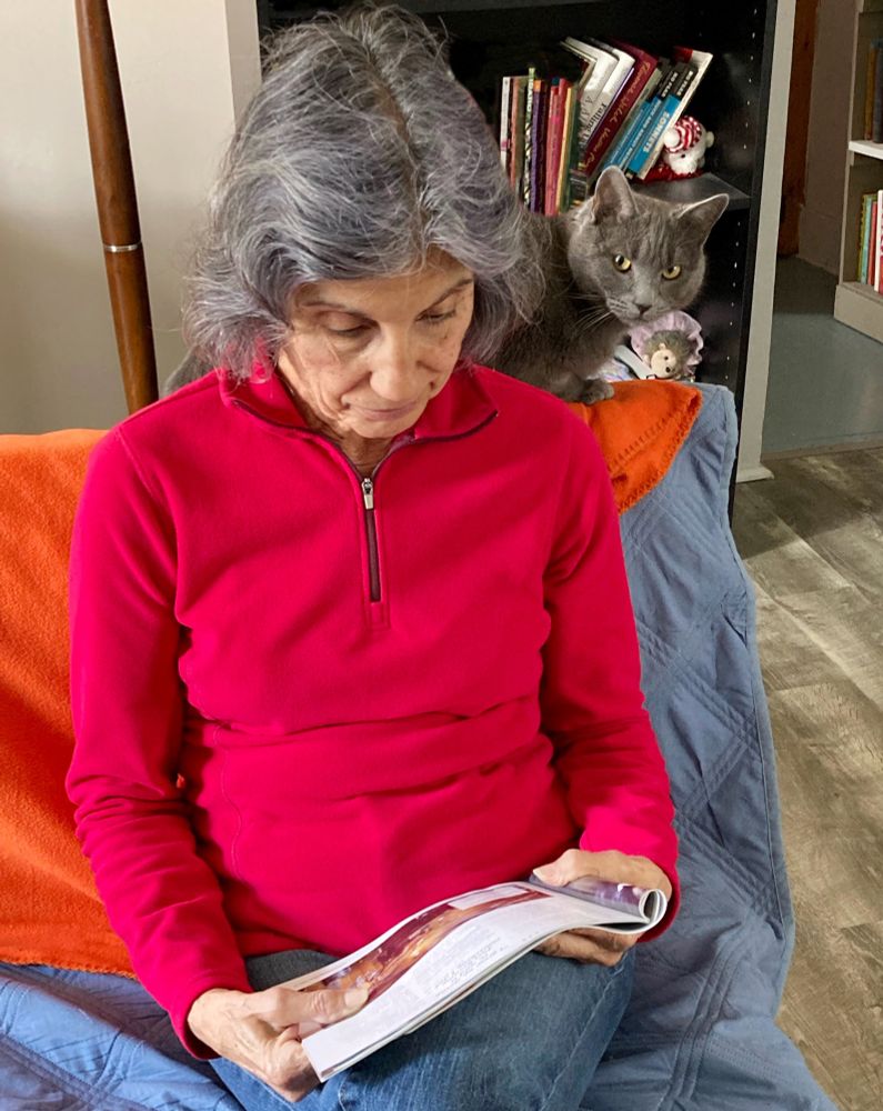 A gray cat, Theo, sits behind her Grandhuman, who reads a magazine on a couch.