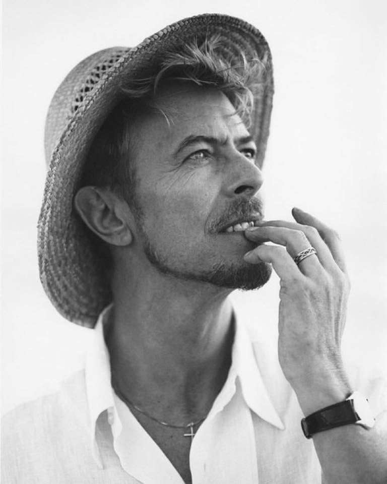 David Bowie wearing a straw hat snd looking up and to the right. His middle fingers held to his lips. He looks eager and yet wistful to me. photo credit Bruce Weber