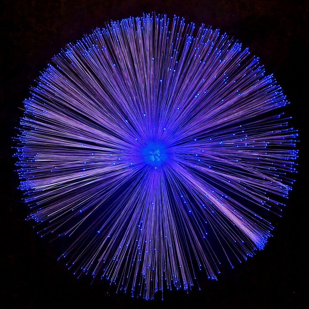 Das Bild zeigt eine kreisförmige Anordnung aus vielen dünnen, lichtleitenden Fasern, die von einem zentralen Punkt ausstrahlen. Die Enden der Fasern leuchten in einem intensiven Blau. Der Hintergrund ist schwarz, wodurch der Kontrast der blauen Lichtpunkte verstärkt wird. Das Gesamtbild wirkt abstrakt und erinnert an einen leuchtenden Feuerwerkskörper oder ein kosmisches Phänomen.
