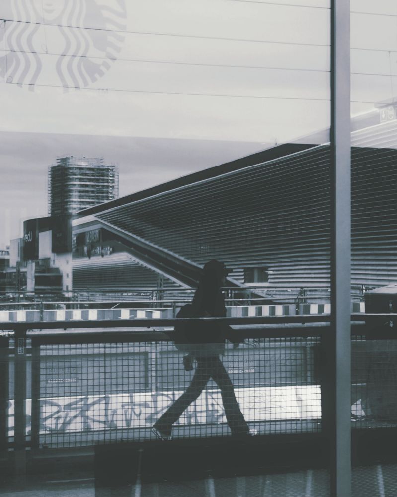 Das Bild zeigt die Silhouette einer Person, die sich im vorbeilaufen in einer Schaufensterscheibe spiegelt. Ausserdem spiegelt sich dort ein Gitterzaun, der mit Graffiti besprüht ist und das moderne Gebäude einer Einkaufsmall. Das Starbucks-Logo ist schwach auf der Scheibe zu sehen. Das Bild ist Schwarz-Weiß. 