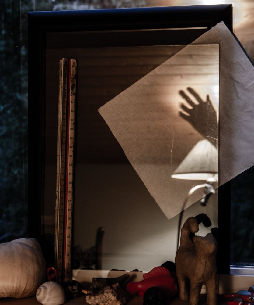 A black picture frame sits within a window. On the ledge in front of it sits a number of sea shells and other small odds and ends including a small papier mache cat.  Within the picture frame is a facial tissue held in place by a small pane of glass which in turn is held in place by a triangular measuring scale.  The transparent glass surface shows part of the tissue and also reflects a lamp in the background and a reflection of a hand.  