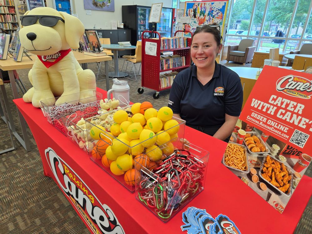 A Raising Cane's employee smiles at a table with various game prizes, including stress relief balls, mini stuffed yellow labs, necklaces, carabiners, and bracelets.