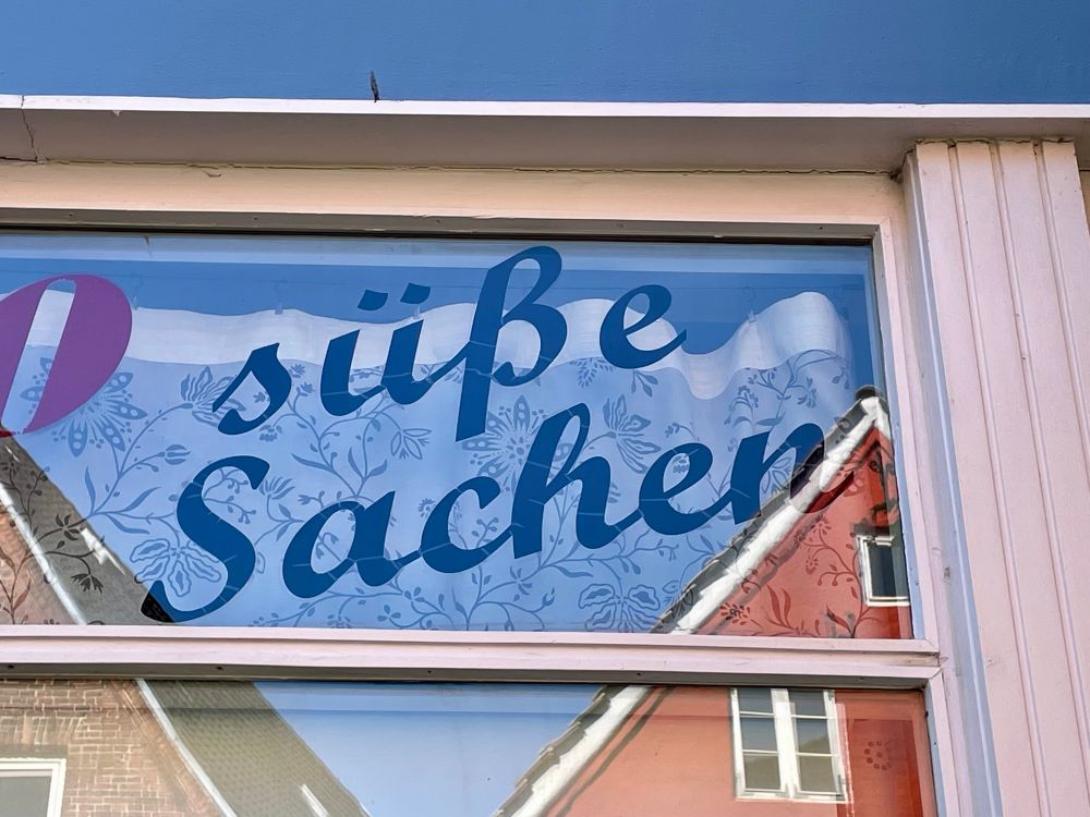 Blaue Schrift ”Süße Sachen” auf einer Schaufenster-Scheibe mit Spiegelung von Hausdächern - Flensburg 