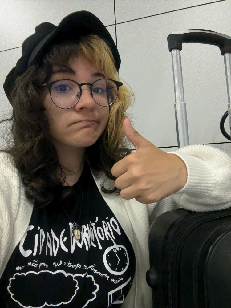 selfie minha (pessoa branca de cabelo castanho escuro e metade da franja loira, estou usando óculos, chapéu preto, uma camisa da banda cidade dormitório e um cardigã branco) dando um joinha ao lado da minha mala