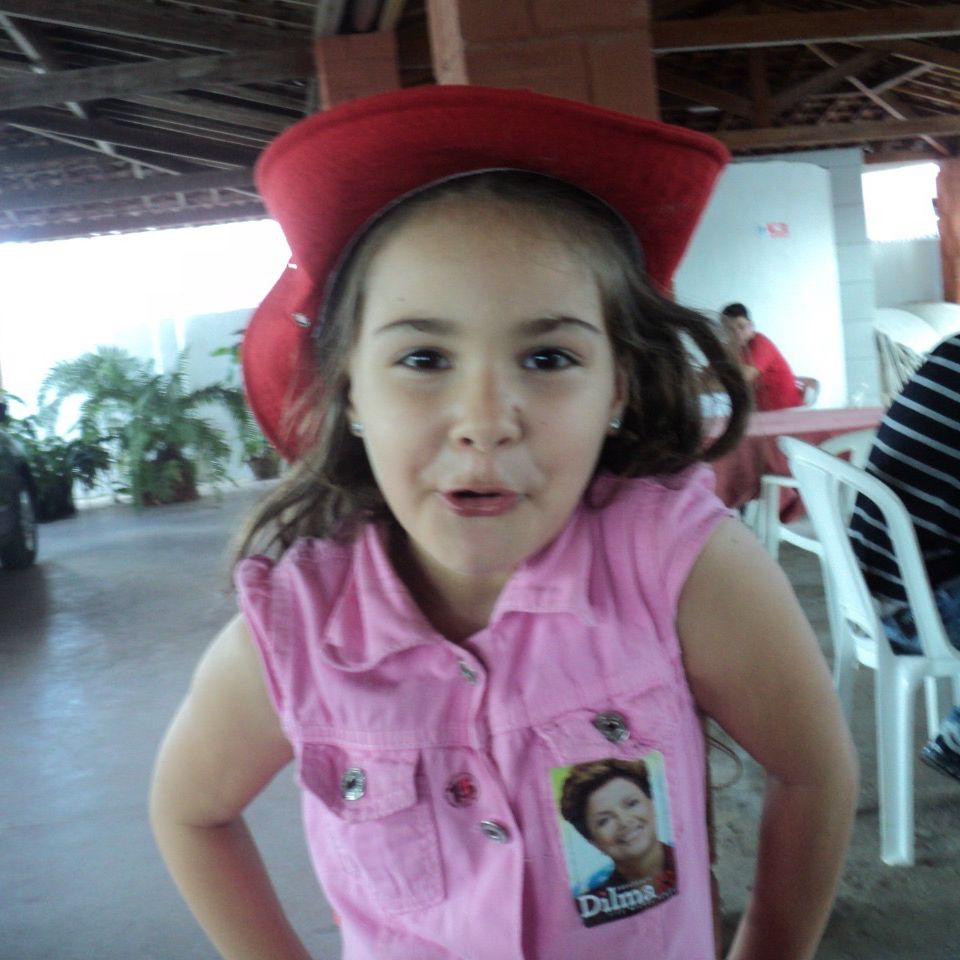 eu criança, cabelo longo castanho, olhos castanhos escuros, branca, devia ter uns 6 anos. estou com um macacão rosa, um chapéu de pescador vermelho, e um adesivo da dilma no peito. também faço um bico com a boca entreaberta e estou com as mãos na cintura.