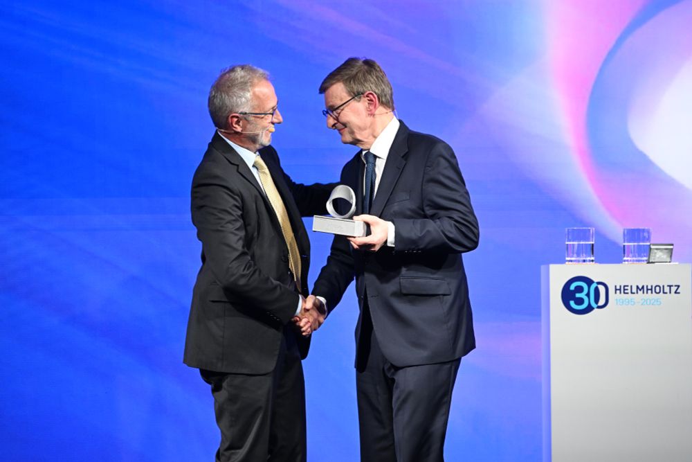 Bei der Amtsübergabe der Helmholtz-Gemeinschaft überreicht Otmar Wiestler (rechts) das Möbiusband an Martin Keller (links). Beide tragen Anzug und Krawatte, stehen auf einer Bühne vor blauem Hintergrund und schütteln sich die Hand. Rechts im Bild ist ein Podest mit dem Helmholtz-Logo und der Aufschrift „30 Helmholtz 1995–2025“ zu sehen. Bild: Oliver Walterscheid