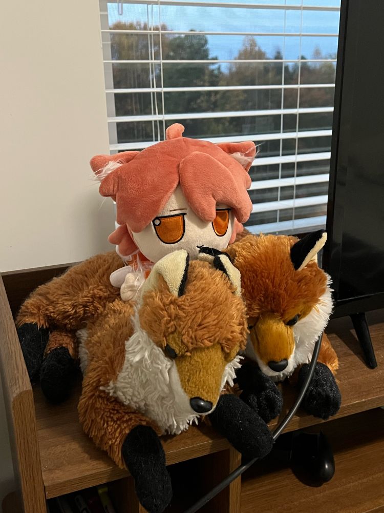 colon plushie sitting on top of 2 fox plushies, window behind