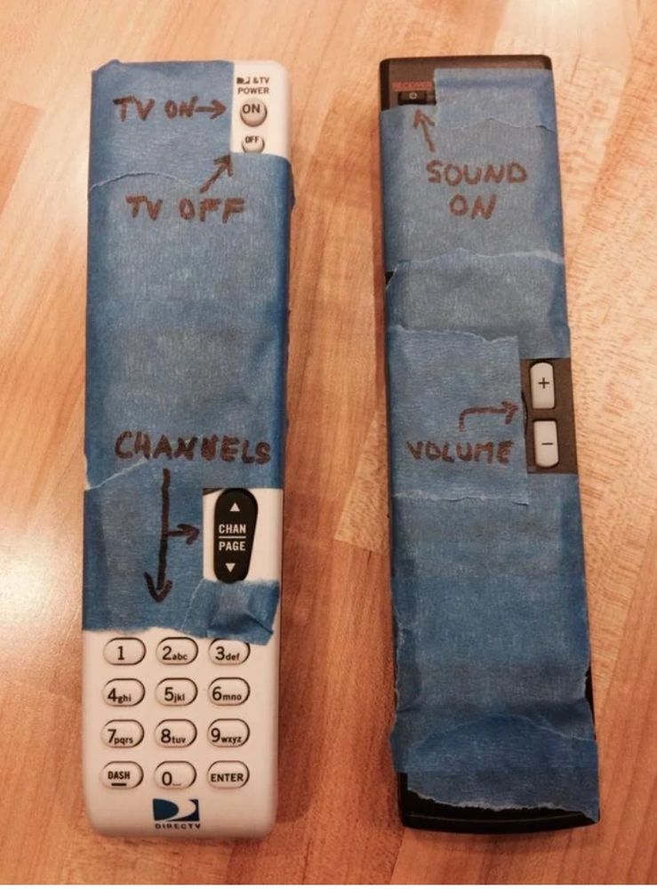 two remote controls for a TV setup with painters tape covering the many superfluous buttons and instructions for power, sound, volume, and channels written in sharpie on the tape.