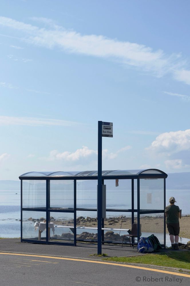 A glass bus shelter with a bus stop in front of it. Inside the shelter is a table with a tablecloth at one end, and a chair at the other. Beside the shelter a man is leaning on it surrounded by luggage and looking out to the sea; in the background is pale blue sky, pale blue sea, and the Scottish mainland in the distance.