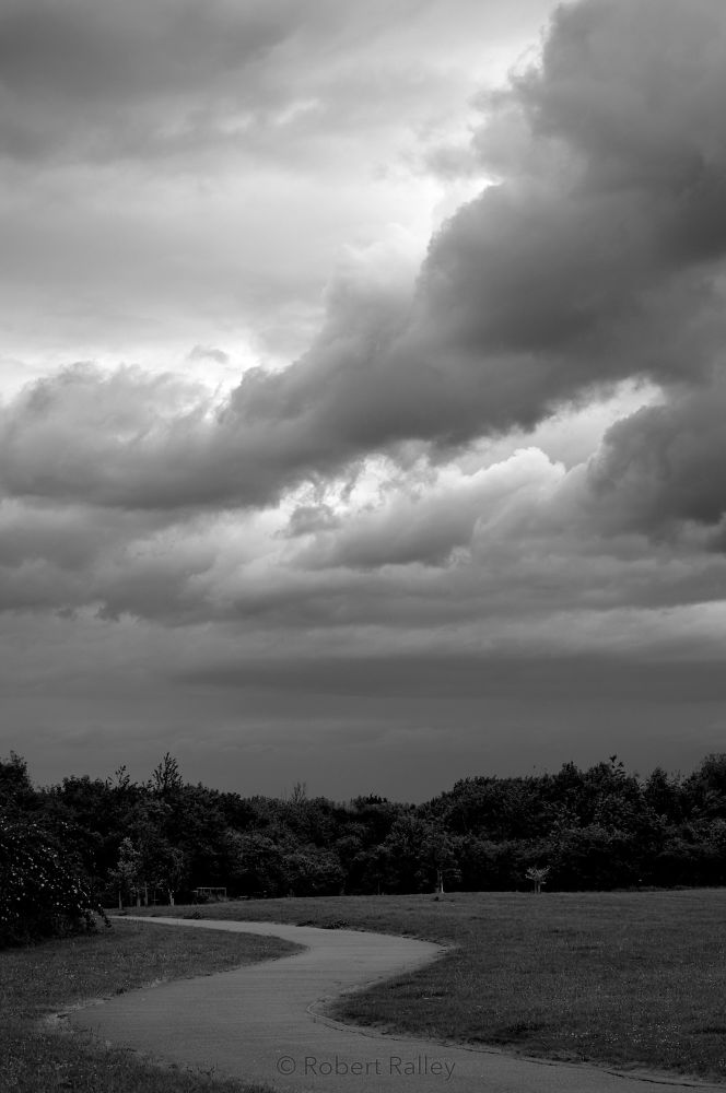 A path winding through grassy parkland towards shrubs and trees. It is gloomy. Overhead there are thick rolls of heavy cloud, crisp-edged, with brighter patches where the sun is shining behind them.