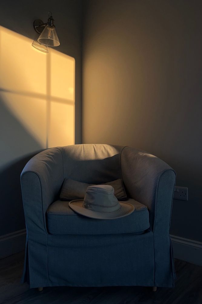 The corner of a white-walled room with an easy chair in it, and a hat sitting on the chair. A golden evening light is shining through a window onto one wall and the top of the chair's back.