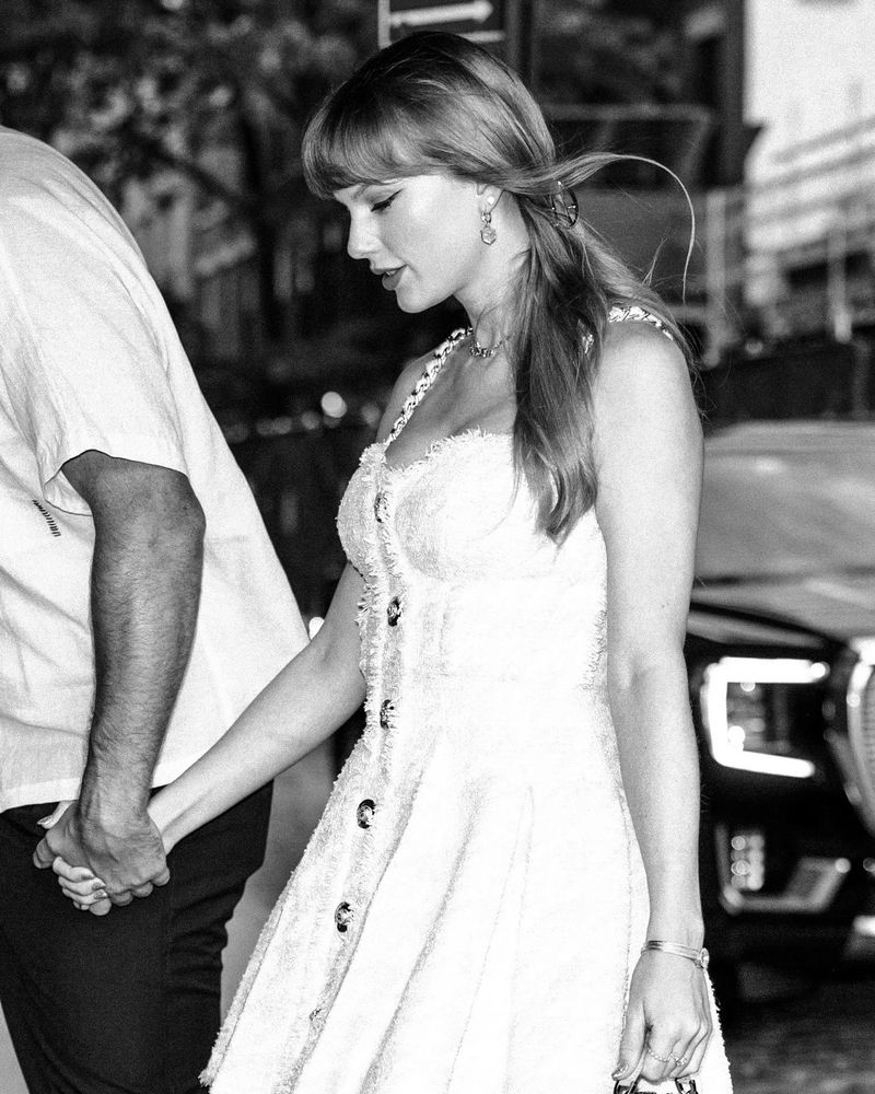 A black and white edit of Taylor Swift out in NYC wearing a tweed light pink short dress with gold hardwire details and her blonde hair is in a half-up, half-down style (June 28, 2025)