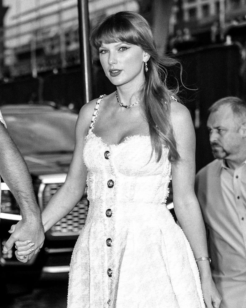 A black and white edit of Taylor Swift out in NYC wearing a tweed light pink short dress with gold hardwire details and her blonde hair is in a half-up, half-down style (June 28, 2025)